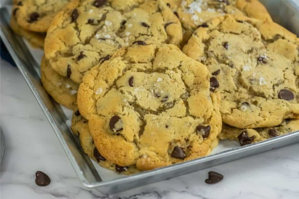 A tray of freshly baked chocolate chip cookies, crafted from a delightfully simple recipe and sprinkled with sea salt, rests invitingly on a marble surface.