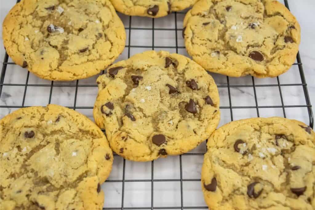 Six freshly baked chocolate chip cookies with sea salt flakes rest on a cooling rack, crafted from a secret recipe that guarantees delight in every bite.