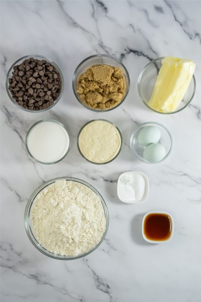 Baking ingredients on a marble surface for a chocolate chip cookies recipe: chocolate chips, brown sugar, butter, sugar, condensed milk, eggs, flour, baking powder, salt, and vanilla extract in glass bowls.