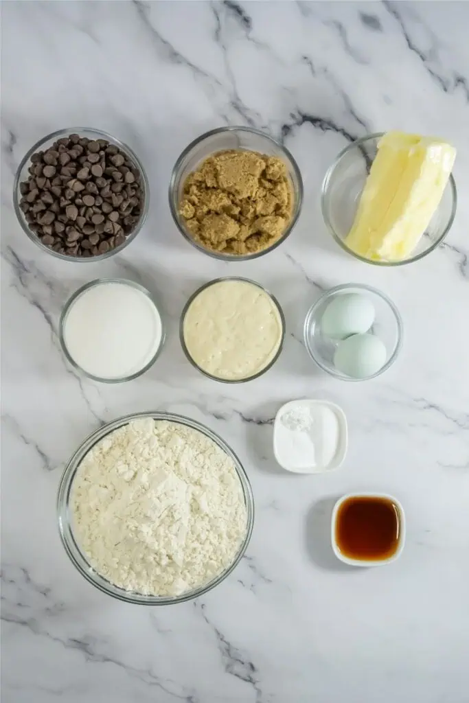 Baking ingredients on a marble surface for a chocolate chip cookies recipe: chocolate chips, brown sugar, butter, sugar, condensed milk, eggs, flour, baking powder, salt, and vanilla extract in glass bowls.