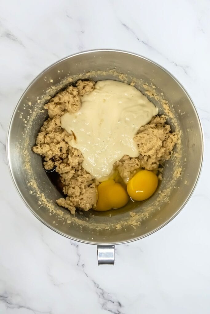 A mixing bowl brimming with chocolate chip cookie dough, accompanied by two eggs and cream cheese, sits invitingly on a marble surface.