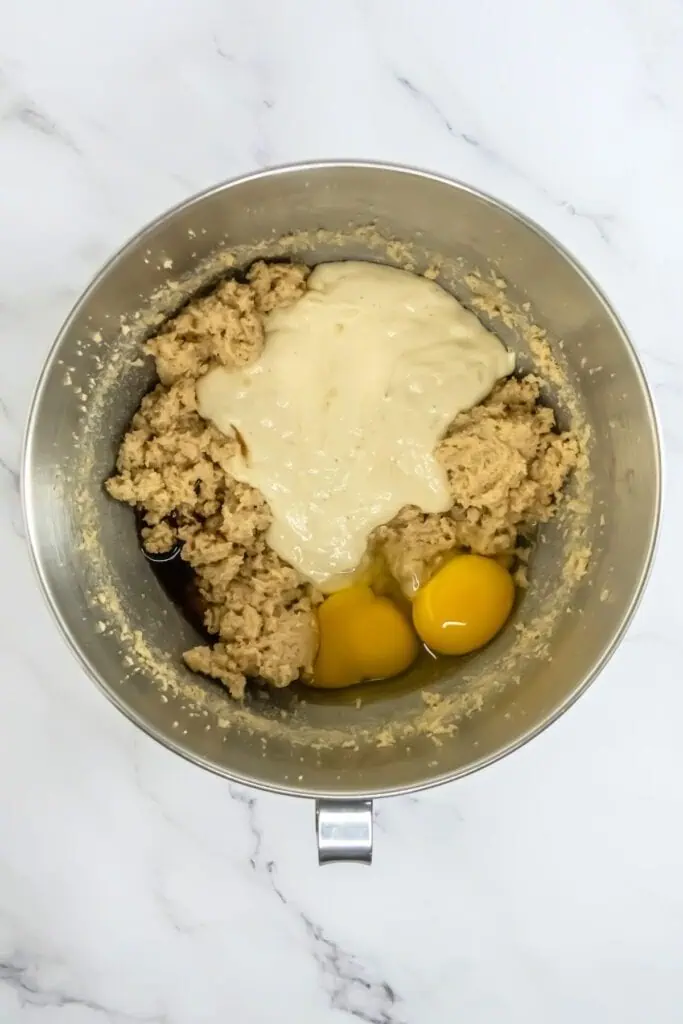 A mixing bowl brimming with chocolate chip cookie dough, accompanied by two eggs and cream cheese, sits invitingly on a marble surface.