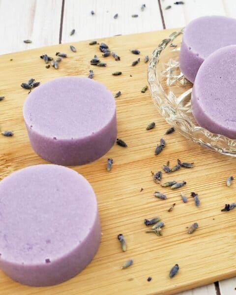 Round lavender soap bars rest on a wooden board, surrounded by scattered dried lavender flowers. These exfoliating bars offer a gentle cleanse, transforming your routine into a soothing ritual.