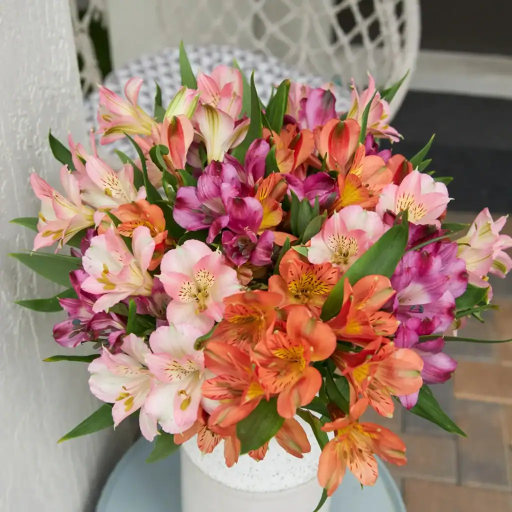 A bouquet of assorted alstroemeria flowers in shades of pink, orange, and purple graces a white vase on a patio, showcasing an elegant touch that's surprisingly affordable. Perfect for any occasion and easily accessible through flower delivery from Walmart.