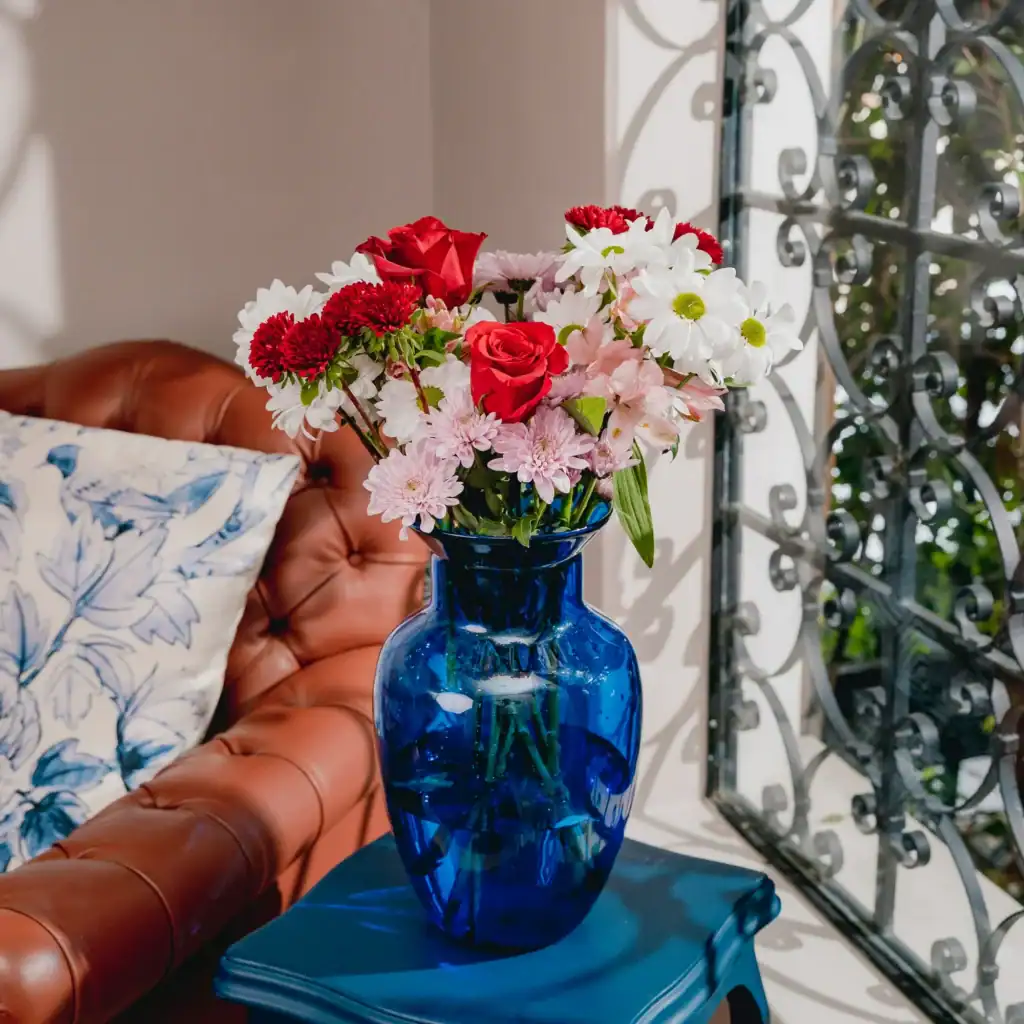 A bouquet of red roses, white daisies, and pink flowers rests in a blue vase on a small table next to a brown sofa and window with decorative ironwork, perfect for those seeking an elegant touch without breaking the bank through flower delivery.