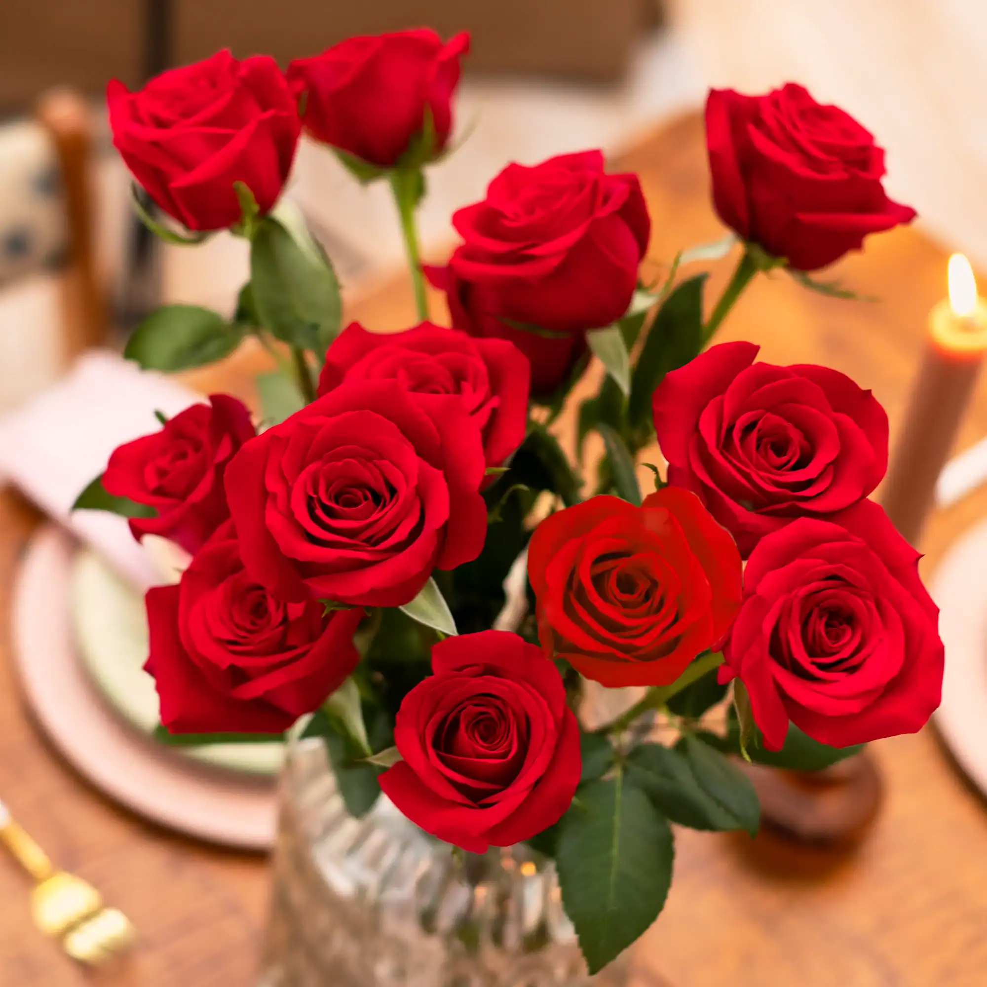 A bouquet of red roses in a glass vase graces a wooden table, alongside a lit candle and blurred tableware. Whether for Walmart shoppers or those seeking cheap flower delivery, this setting adds elegance to any occasion.