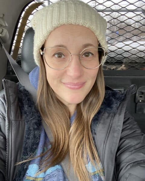 Person wearing a white beanie and glasses, sitting in a car with a metal grid behind them.