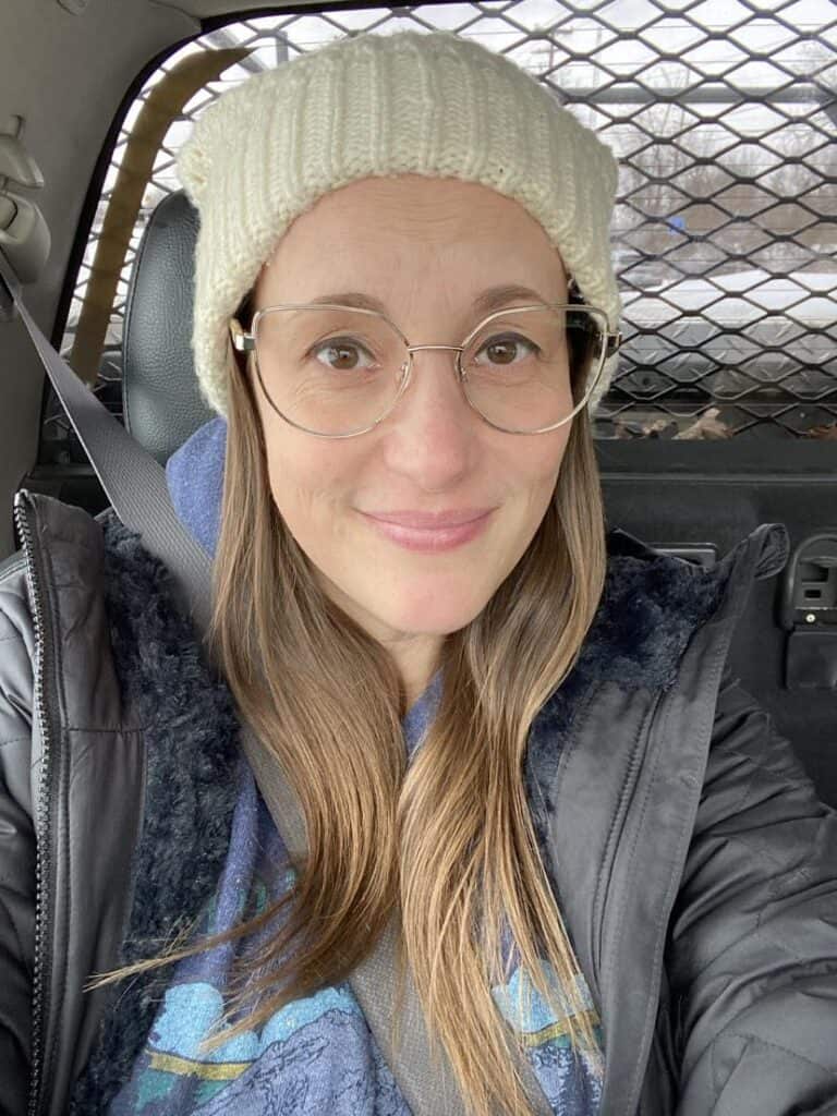 Person wearing a white beanie and glasses, sitting in a car with a metal grid behind them.