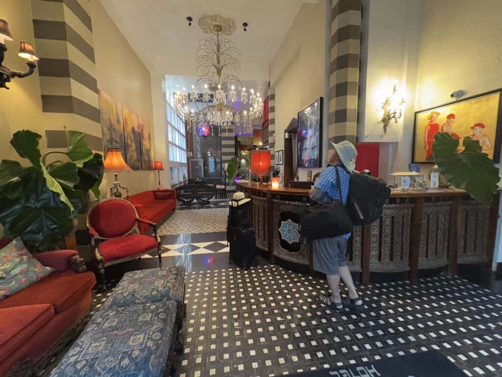 A person with luggage stands at a vintage hotel reception. Ornate chandelier, large plants, and red upholstered chairs decorate the space.