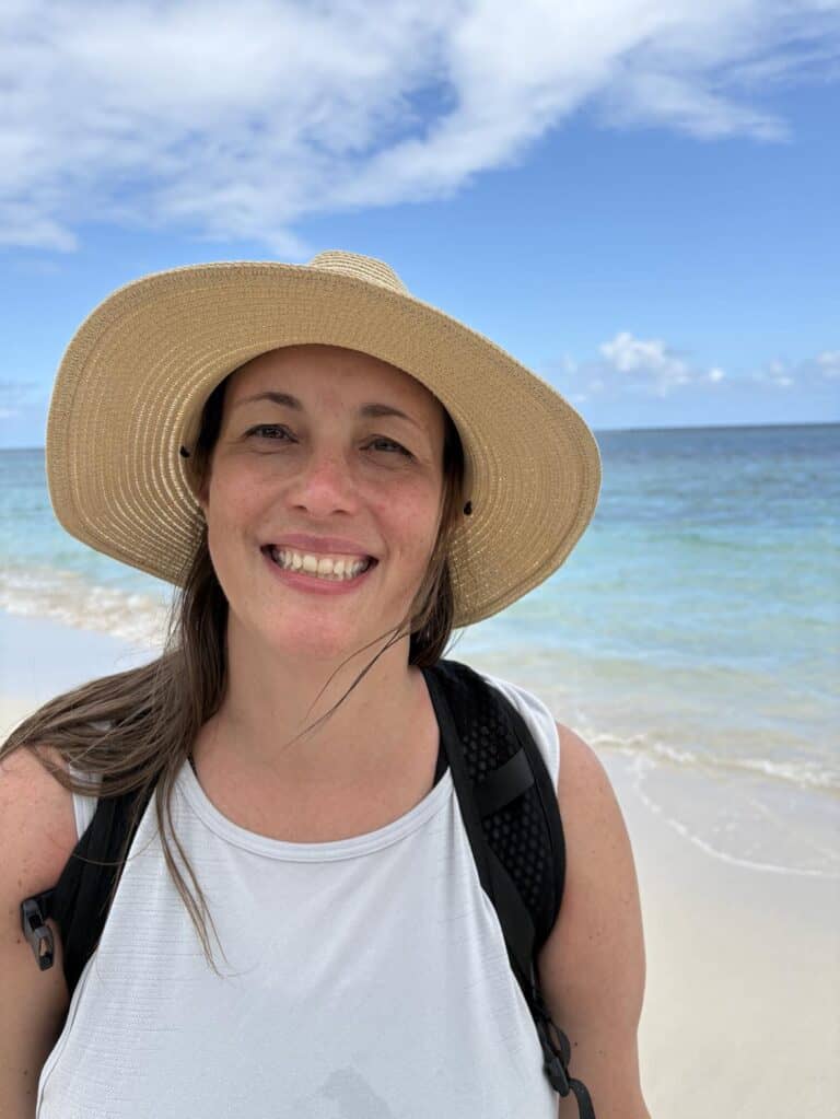 A person wearing a straw hat and a white shirt smiles while standing on a beach with the ocean and blue sky in the background.