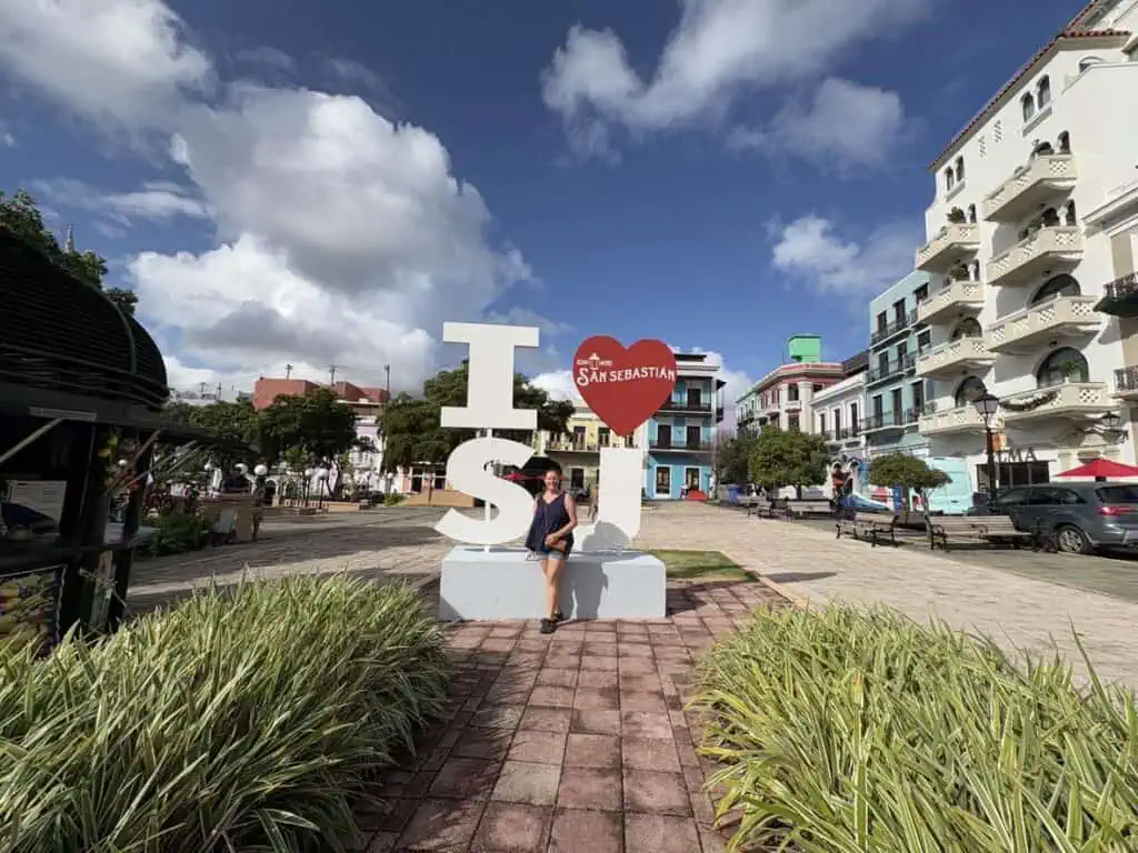 A person sitting on a large "I ❤️ SJ" sculpture in a plaza with colorful buildings, under a partly cloudy sky.