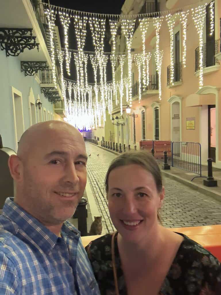 A man and woman smiling in a cobblestone street at night; decorative lights hang overhead, and brightly colored buildings line the street.