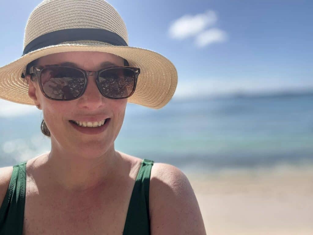 Person wearing a sun hat and sunglasses, smiling at the beach with clear blue sky and ocean in the background.