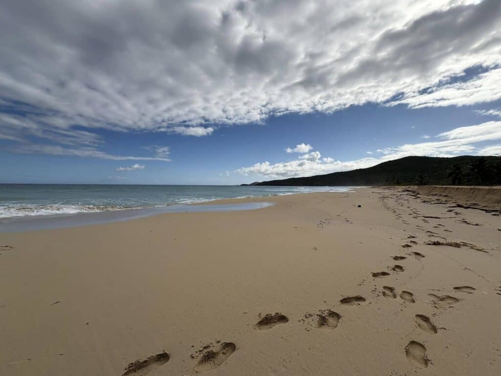 A sandy beach with footprints leading towards the sea, under a partly cloudy sky. Hills are visible in the distance.