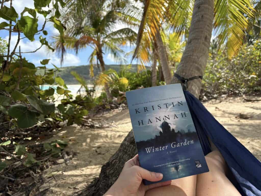 Person holding the book "Winter Garden" by Kristin Hannah while lying in a hammock on a tropical beach with palm trees.