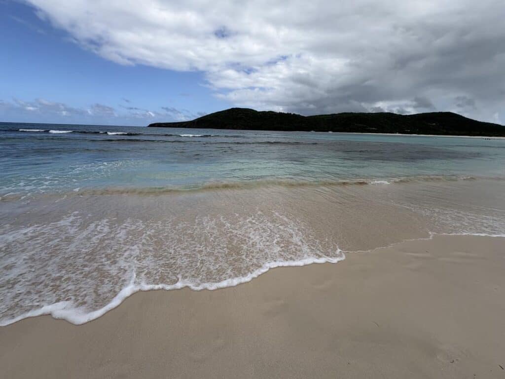 Sandy beach with gentle waves lapping at the shore. The ocean is light blue and green, under a partly cloudy sky with a green hill in the distance.