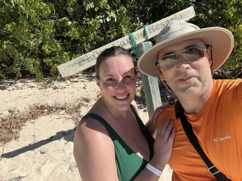 A man and woman smiling on a sandy beach with greenery in the background. The man wears a sun hat and sunglasses, while the woman is in a green swimsuit.