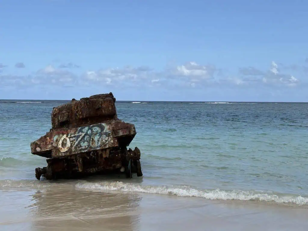 Rusty shipwreck with faded numbers on sandy beach, partially submerged in clear ocean water under a blue sky with scattered clouds.
