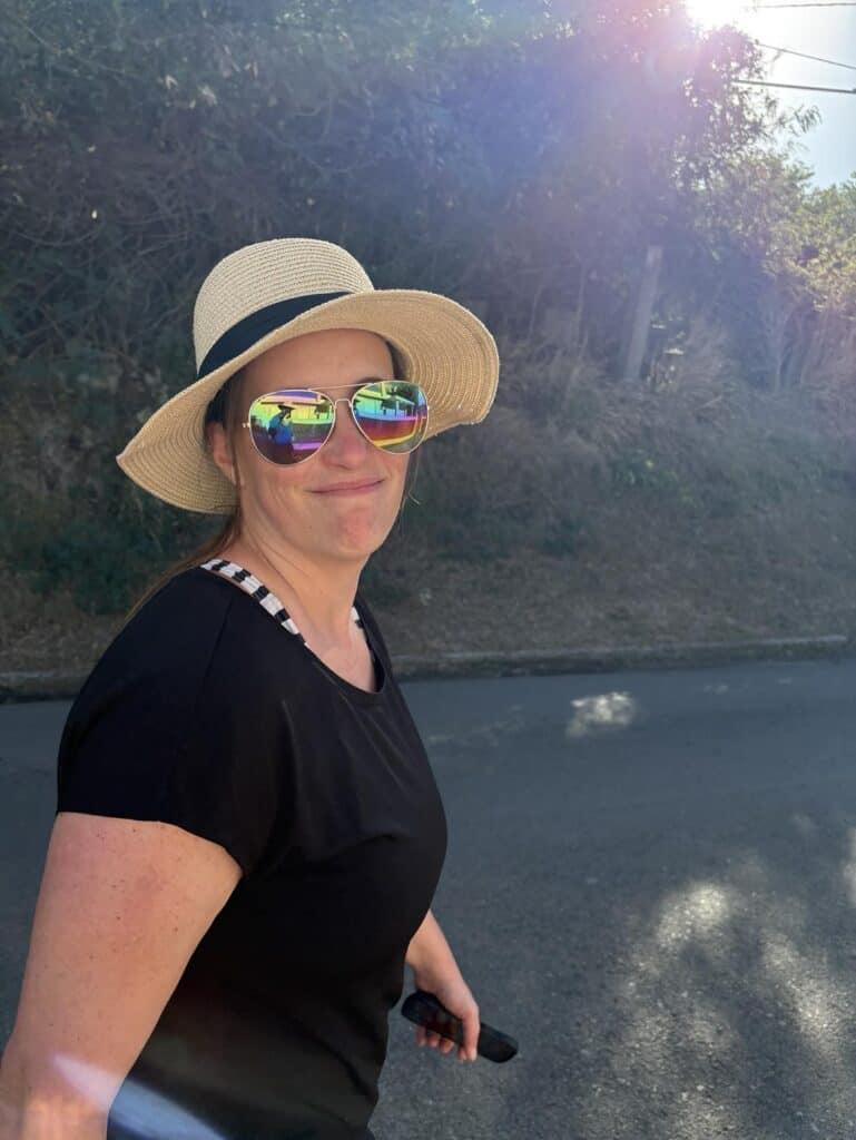 A woman wearing a straw hat and reflective sunglasses stands on a sunlit road, exuding post-vacation blues. Dressed in a black shirt, she holds an object in her hand as trees whisper stories of new beginnings in the background.