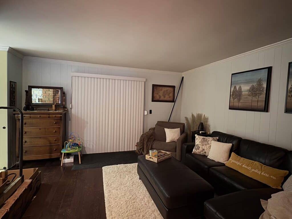 A living room with a black leather sectional sofa, a wooden dresser with a TV, a toy on the floor, and framed art on the walls. A white rug and vertical blinds on the sliding glass door.