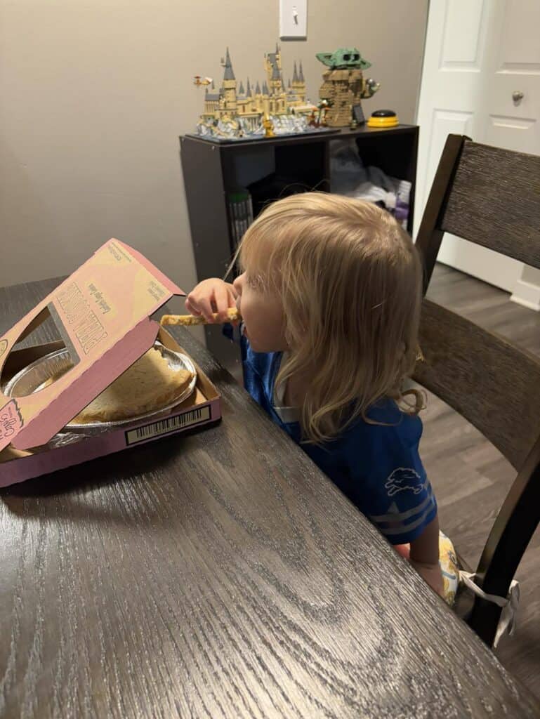 A child in a blue shirt eats pizza from a box at a wooden table. In the background are toy sets on a shelf.