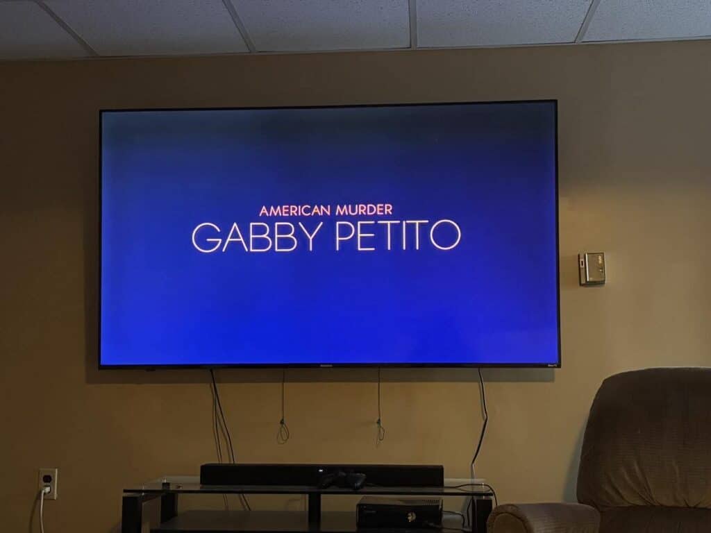 A TV screen displaying the title "American Murder: Gabby Petito" on a blue background is mounted on a beige wall above a stand and recliner chair, inviting moments that feel like the gift of time.