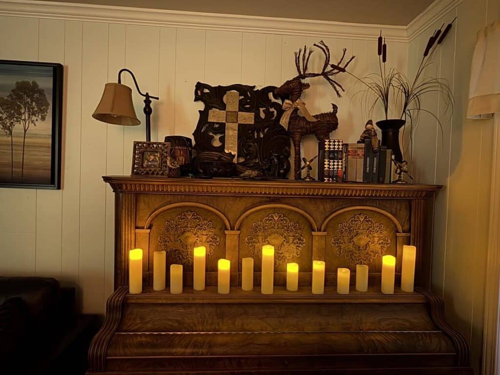 An ornate piano decorated with glowing candles, books, and rustic ornaments, including a cross and reindeer figures, against a wood-paneled wall with artwork and a lamp.