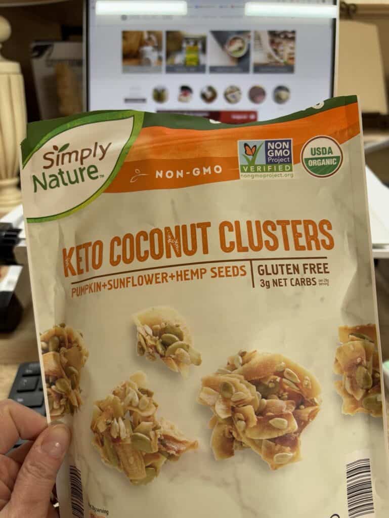 A hand personally holds a bag of Simply Nature Keto Coconut Clusters, featuring ingredients like pumpkin, sunflower, and hemp seeds. In the background, a laptop is open to a recipe post on a website.