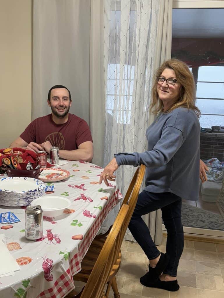 At a dining table adorned with a red and white lobster-patterned tablecloth, two people share a delightful moment. The woman stands, smiling warmly as the man sits comfortably. Drinks and snacks are scattered across the scene, perfect for capturing in a personal post.