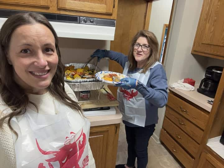 Two women in a kitchen personally bond over their love for seafood. One holds a tray of freshly cooked delicacies; the other presents a plate, both wearing aprons adorned with charming crab designs. The cozy kitchen features wooden cabinets and a trusty coffee maker, setting the perfect scene.