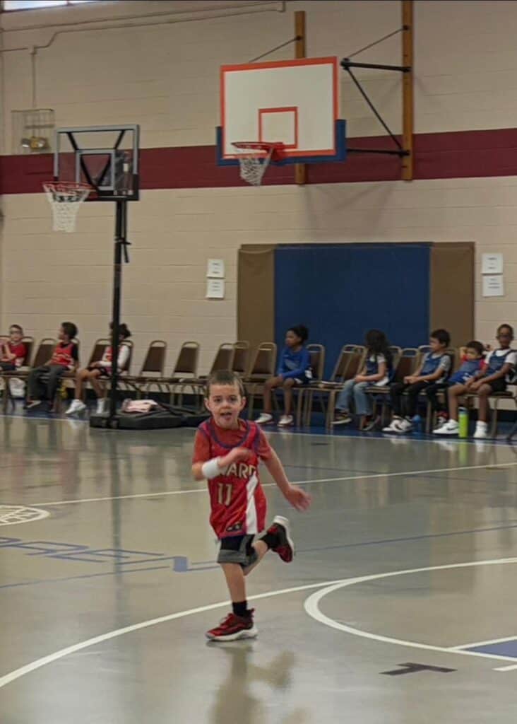 In a charming personal post, a young child in a red basketball jersey dashes across an indoor court, with chairs and spectators gathered near the hoop in the background.