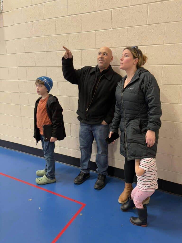 In a vibrant gym, two adults and two children gather. One adult gestures upwards with enthusiasm. The younger child gazes curiously to the side, while the older one looks intently ahead. This scene illustrates a moment where more detailed information would enhance understanding and identify effective keywords.