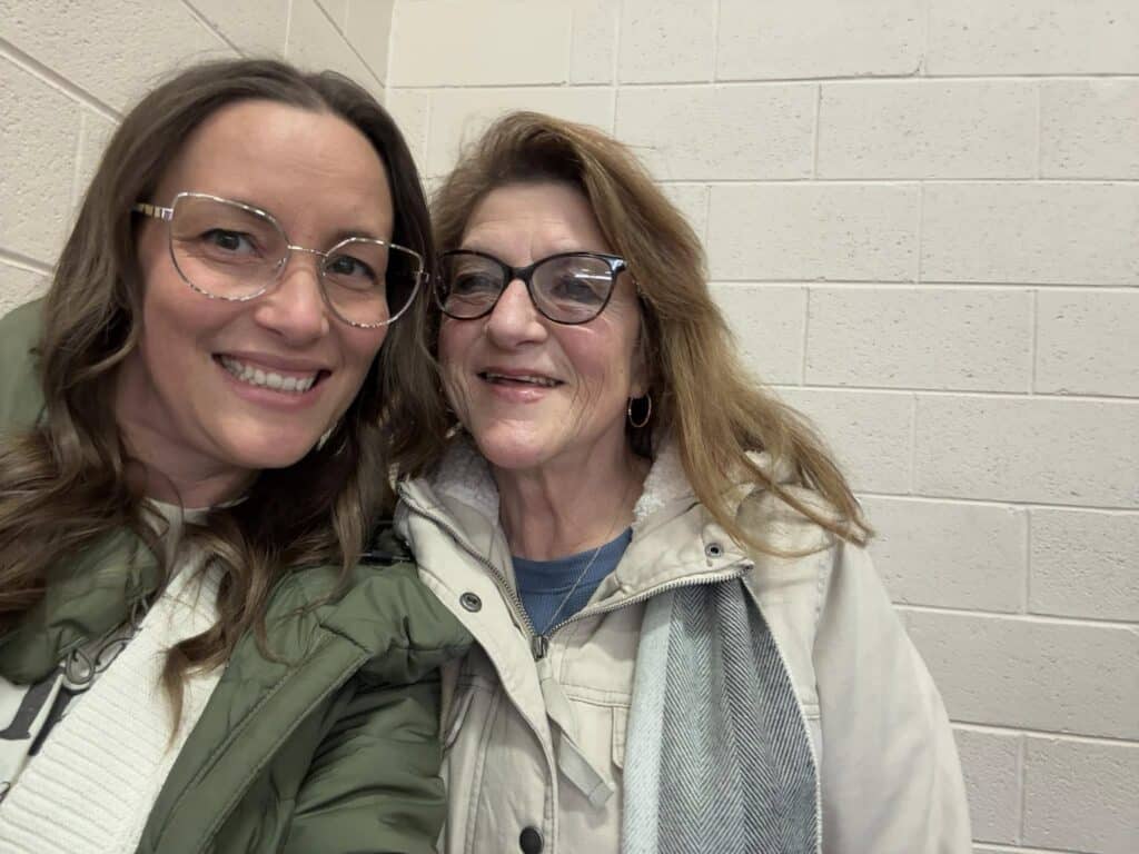 Two women wearing glasses smile for a personal selfie. One sports long brown hair with a green jacket, while the other has sandy hair in a cream jacket. They stand in front of a white brick wall, capturing the moment to post later.