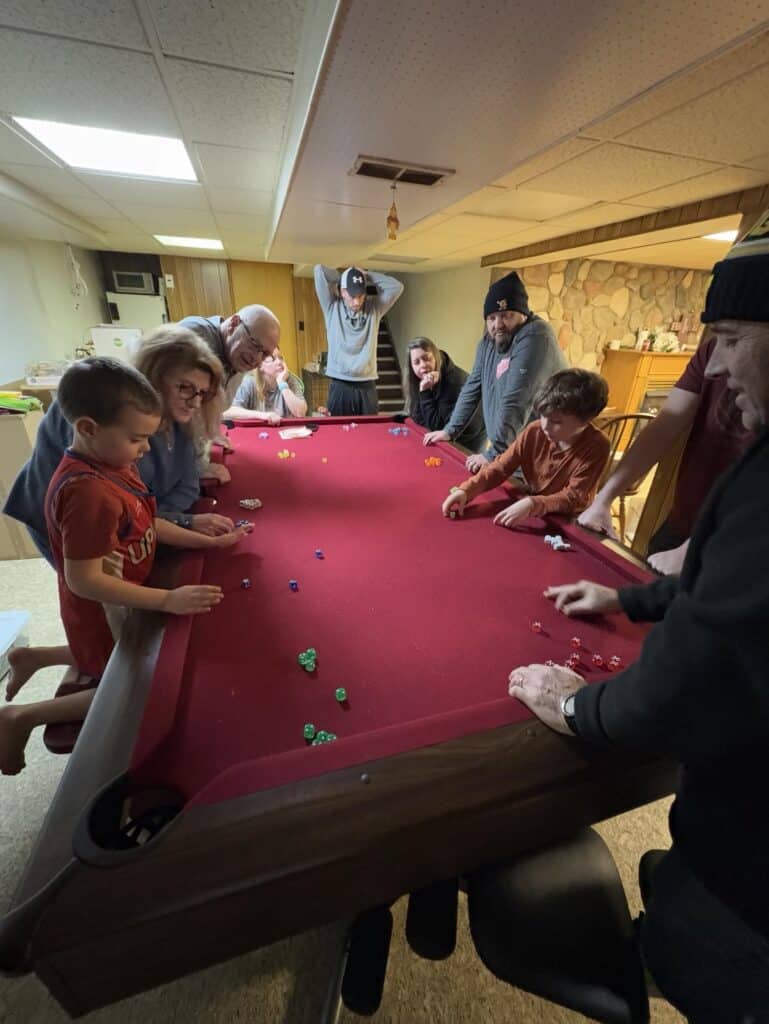 A group of people gathered around a red felt table, playing a dice game in a cozy basement setting. The atmosphere harkens back to the cherished Gift of Time, reminding us how much friendships have changed over the years.