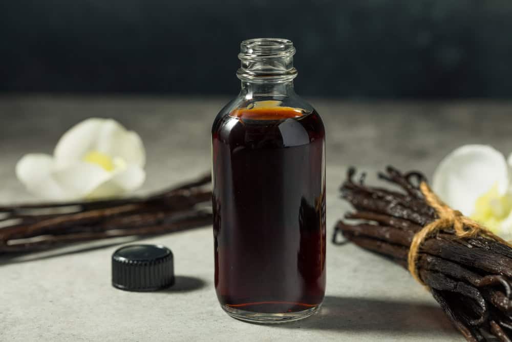 A glass bottle filled with dark liquid and an uncapped lid beside it, reminiscent of a baking essential. Bundles of vanilla pods and white flowers are artfully arranged in the background on a gray surface, perfect for any sweet substitutions.