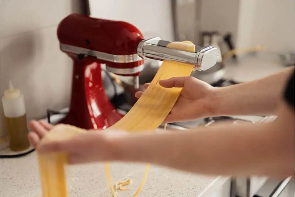 A person expertly using a red stand mixer with a pasta attachment to roll out fresh homemade pasta dough on the kitchen counter, perfecting their favorite pasta recipe.