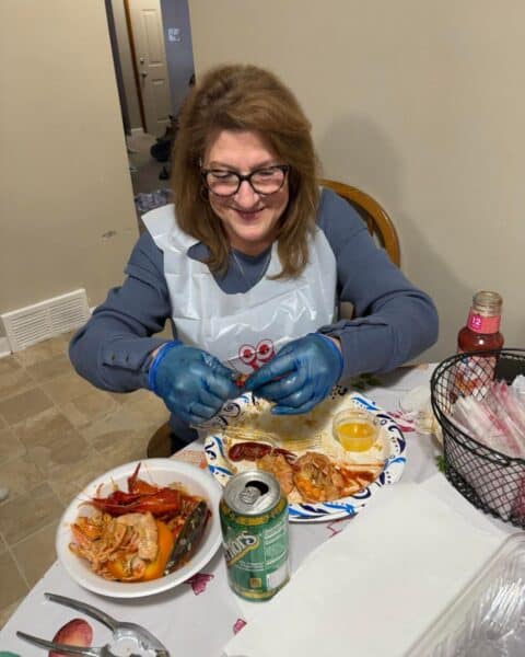 Wearing gloves, a person savors seafood at the table, enjoying shellfish alongside a drink can and a basket of utensils. This meal is more than just food; it's a gift of time, reminiscent of their cherished relationship with Mom.