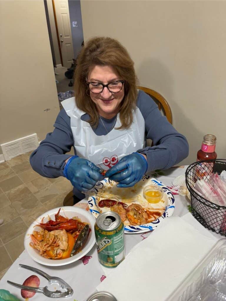 Wearing gloves, a person savors seafood at the table, enjoying shellfish alongside a drink can and a basket of utensils. This meal is more than just food; it's a gift of time, reminiscent of their cherished relationship with Mom.