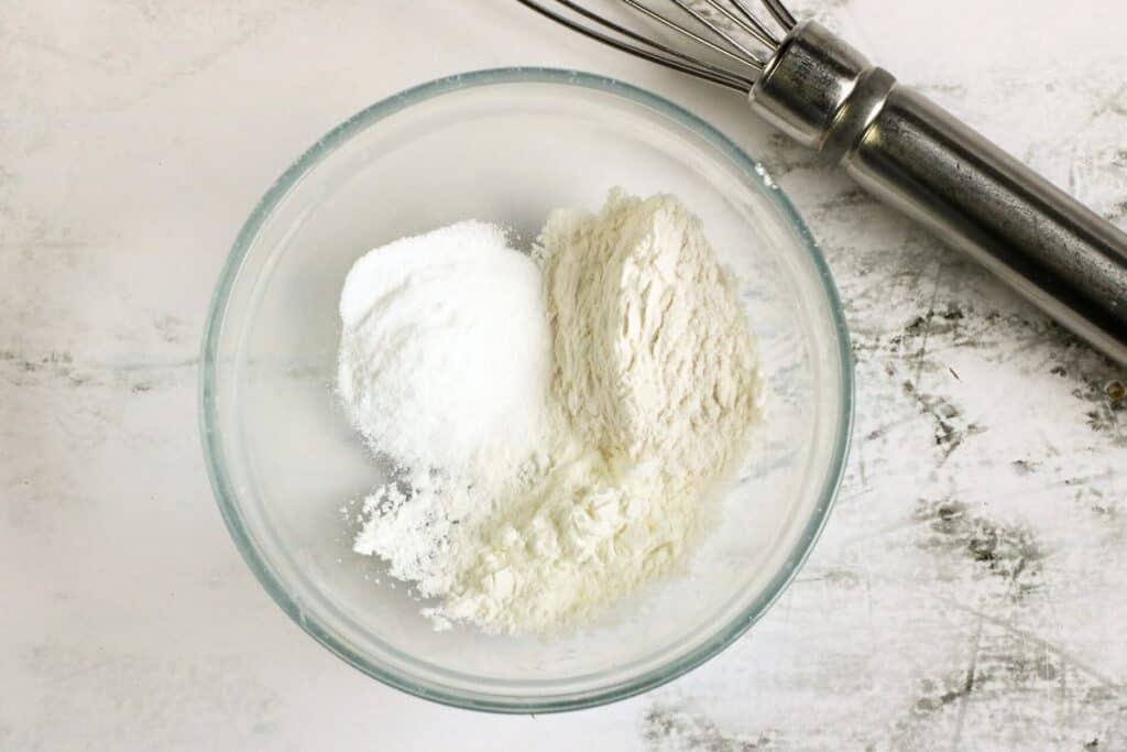 A glass bowl with three white powdery ingredients, essential for any homemade recipe, sits next to a metal whisk on a pristine white surface.