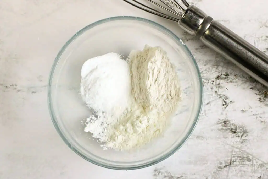 A glass bowl with three white powdery ingredients, essential for any homemade recipe, sits next to a metal whisk on a pristine white surface.
