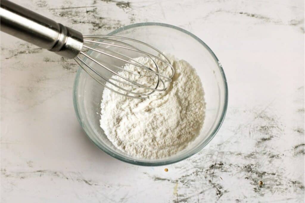 A metal whisk rests in a glass bowl filled with white flour, ready for your homemade baking powder recipe, on a textured white surface.