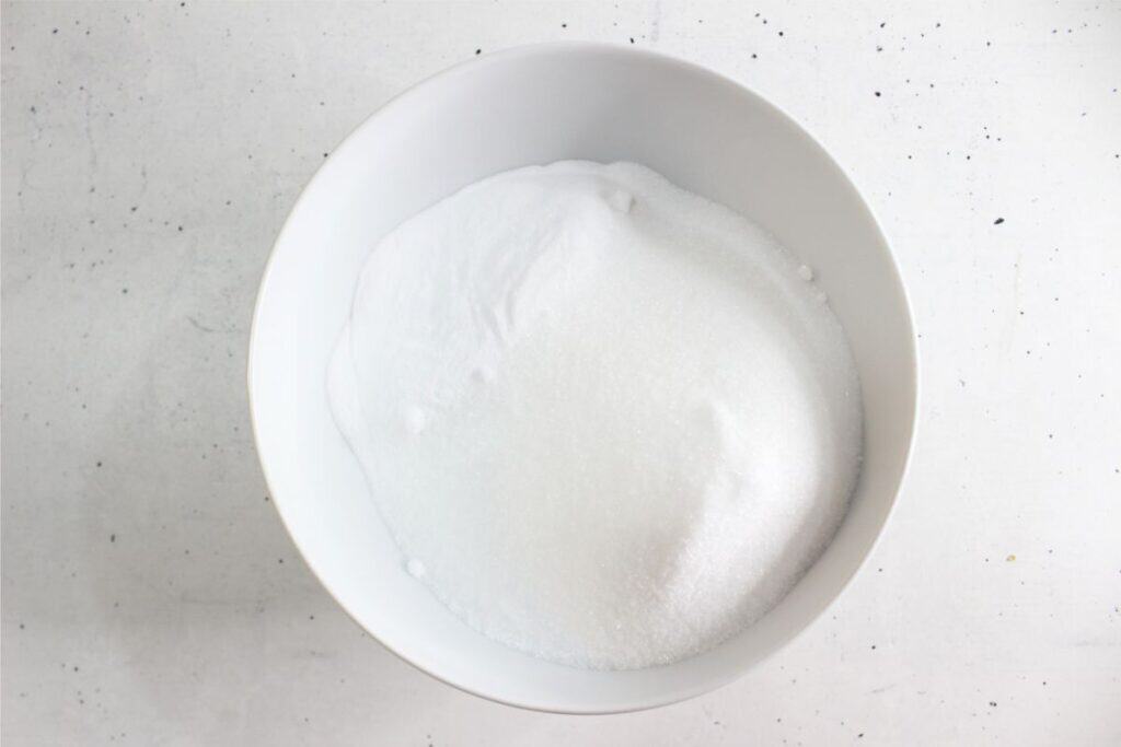 A white bowl filled with granulated white sugar on a white speckled surface, reminiscent of the ingredients for an easy homemade toilet cleaner.