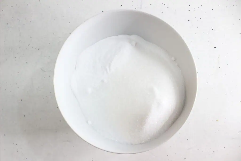 A white bowl filled with granulated white sugar on a white speckled surface, reminiscent of the ingredients for an easy homemade toilet cleaner.