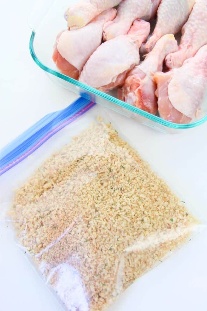 A glass dish holds raw chicken drumsticks beside a plastic bag brimming with homemade shake and bake breadcrumbs.