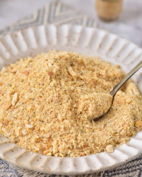 A white plate filled with homemade breadcrumbs, with a spoon partially submerged, rests on a textured cloth.