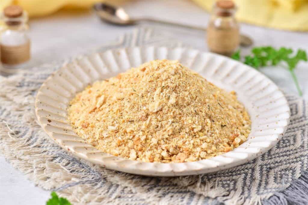 A plate brimming with homemade light brown breadcrumbs, nestled on a textured cloth, while small jars and fresh green herbs add charm in the background.