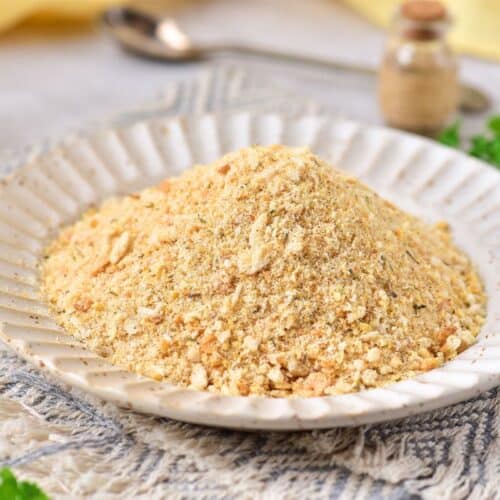 A plate brimming with homemade light brown breadcrumbs, nestled on a textured cloth, while small jars and fresh green herbs add charm in the background.