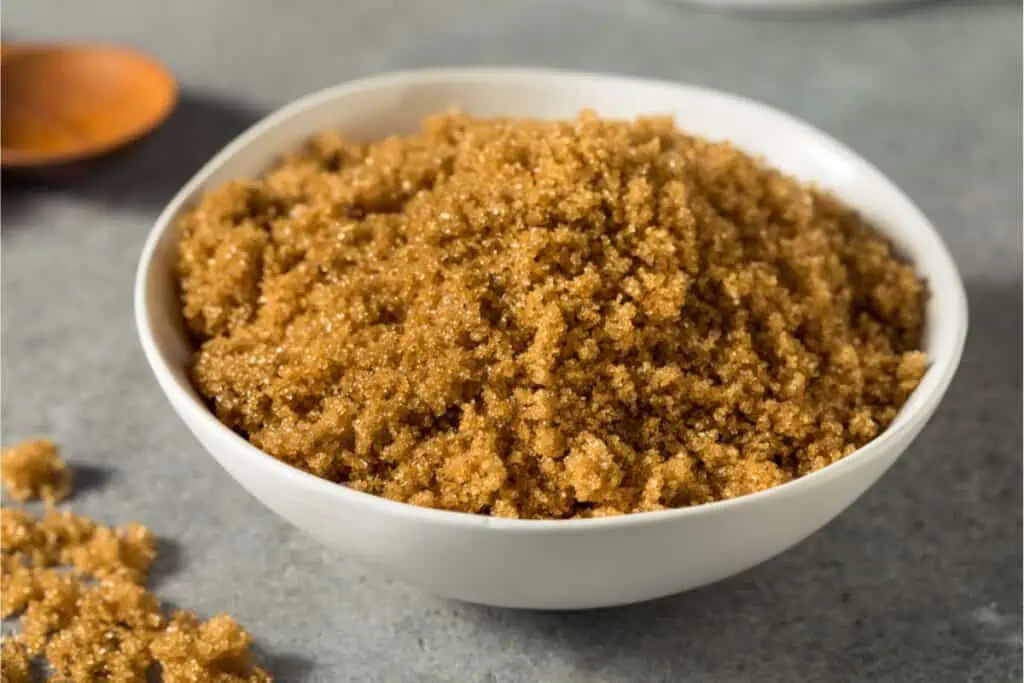 A white bowl filled with brown sugar, perfect for baking substitutions, sits on a gray surface, with a few sugar granules scattered nearby.