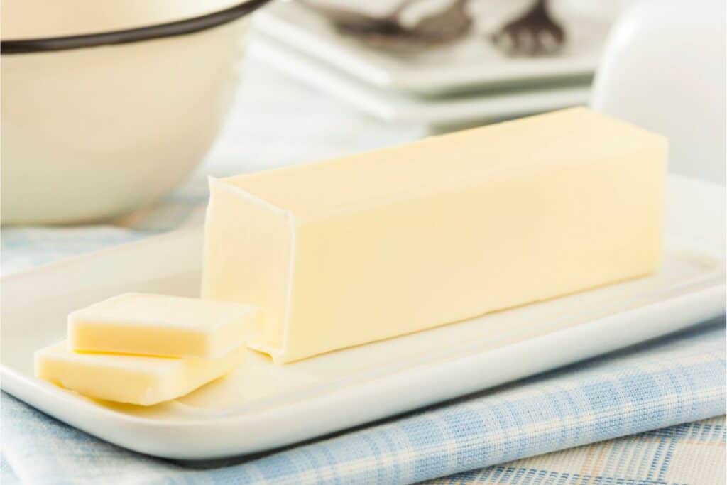 A stick of butter with two slices cut off rests on a ceramic dish, perfect for baking, atop a plaid cloth.