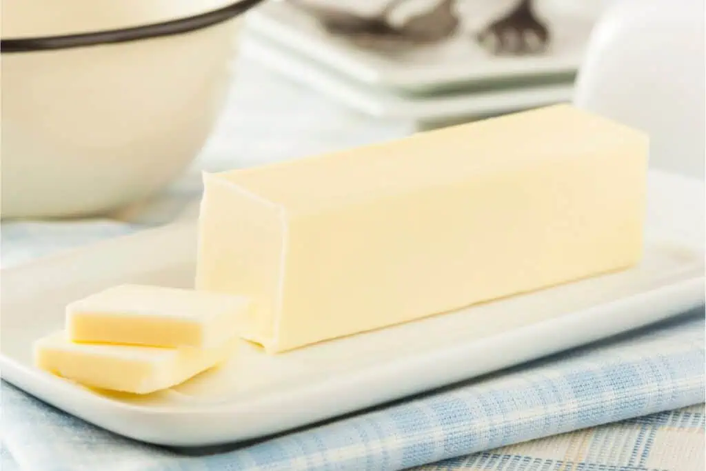 A stick of butter with two slices cut off rests on a ceramic dish, perfect for baking, atop a plaid cloth.